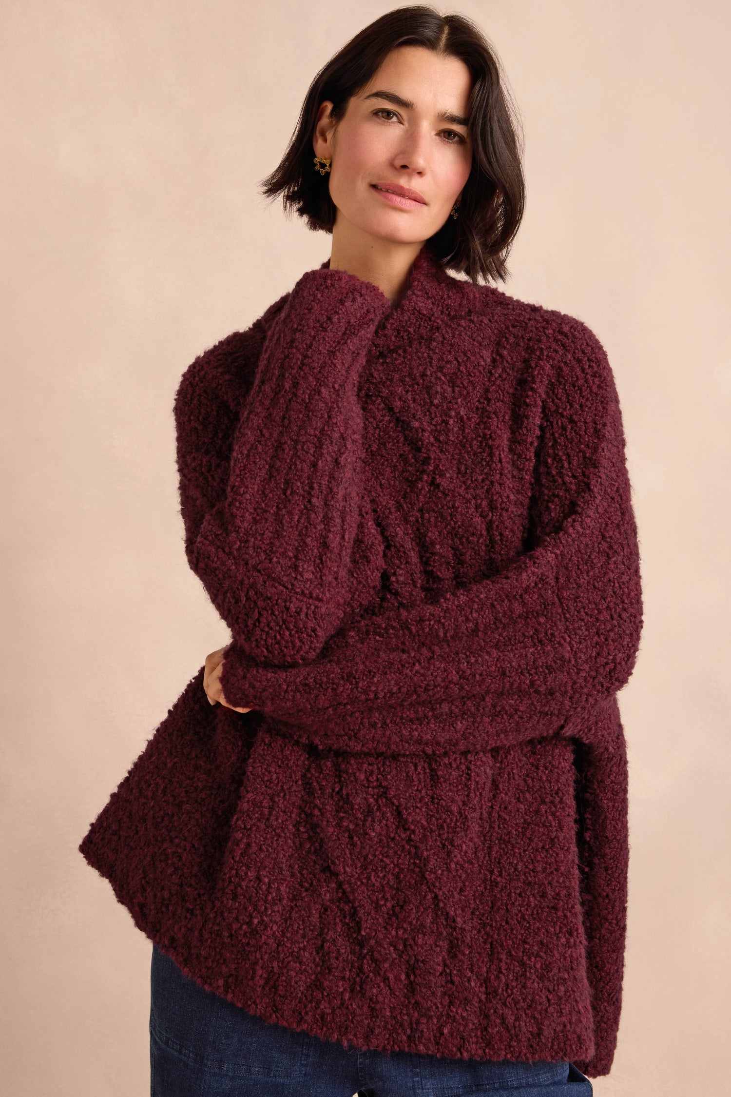 A woman wearing the Bordeaux Boucle Cable Knit Jumper and blue jeans stands against a light beige background with a neutral expression.