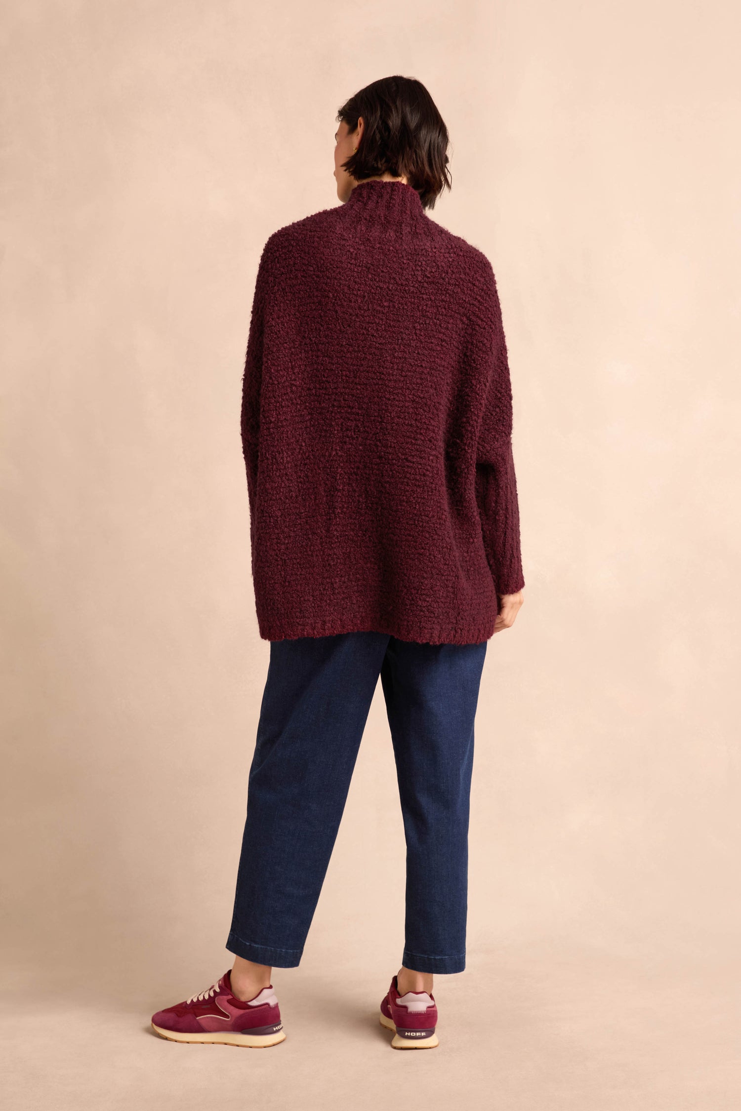 A person stands facing away against a beige background, wearing the Bordeaux Boucle Cable Knit Jumper—crafted in Italy for style and comfort—paired with blue jeans and maroon sneakers. The textured maroon knit elevates this winter look.