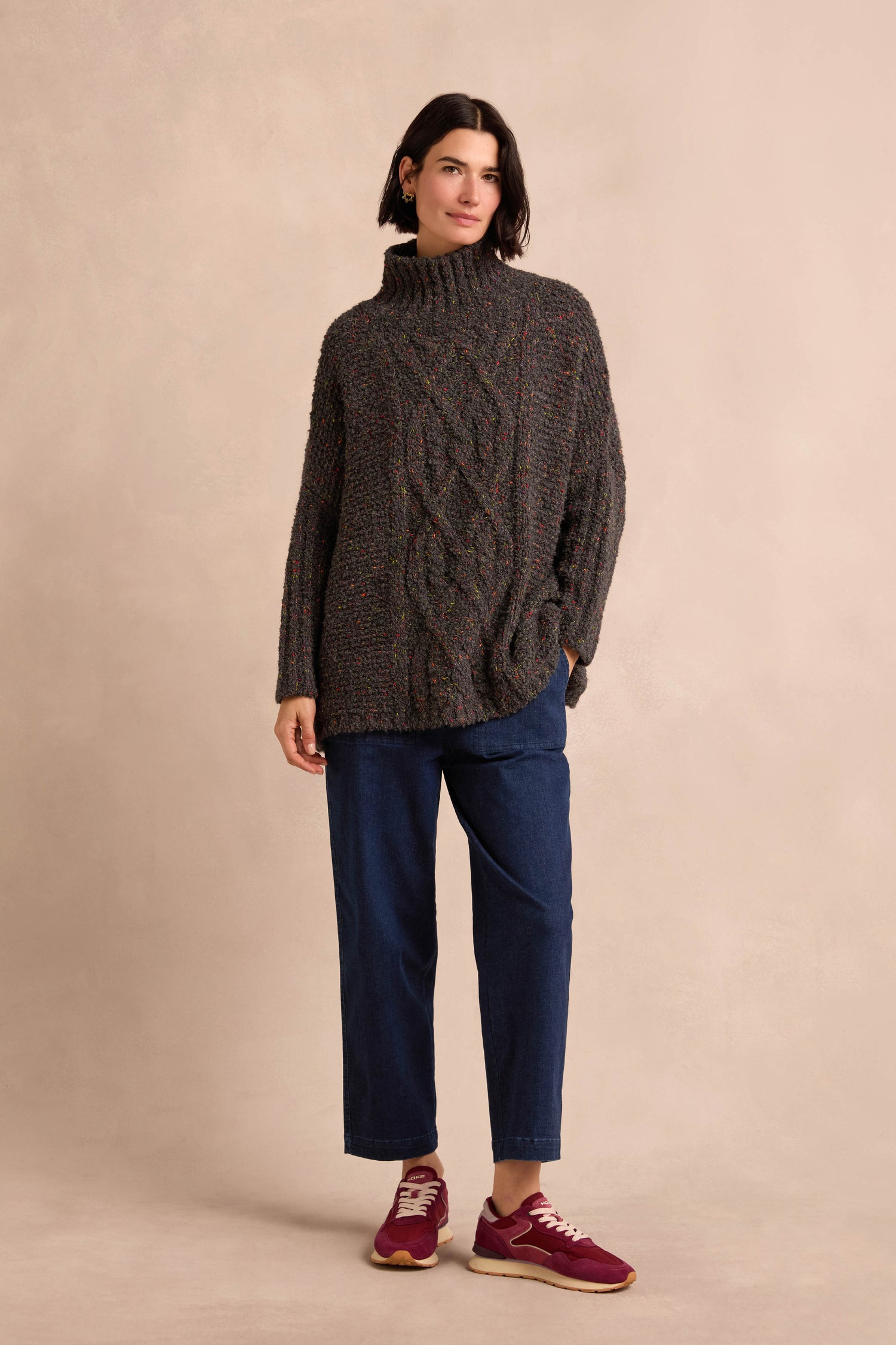 A woman stands against a light background, wearing the Graphite Boucle Cable Knit Jumper—an Italian-made, soft-textured chunky gray turtleneck sweater—paired with blue jeans and red sneakers.