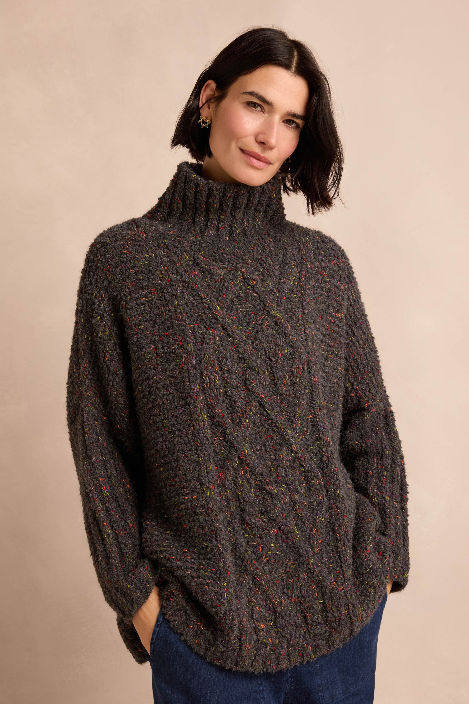 A woman with short dark hair wears the Graphite Boucle Cable Knit Jumper—a soft, textured chunky gray turtleneck sweater—paired with blue pants, standing against a beige background with her hands in her pockets.