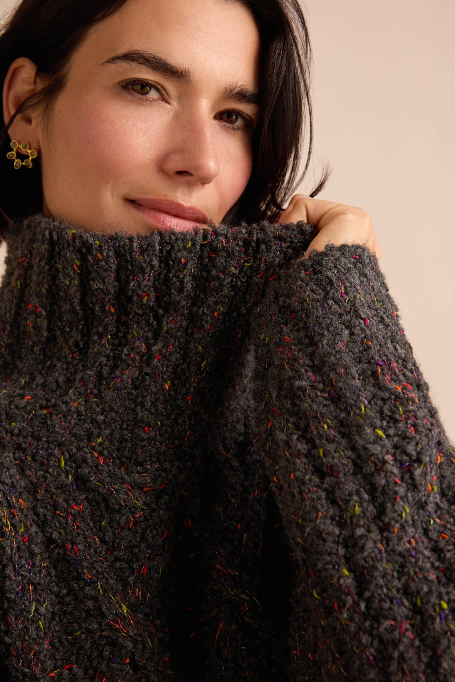 A person with short dark hair wears the Graphite Boucle Cable Knit Jumper, a chunky dark gray sweater with colorful specks and a soft textured finish, holding the collar up to their face.