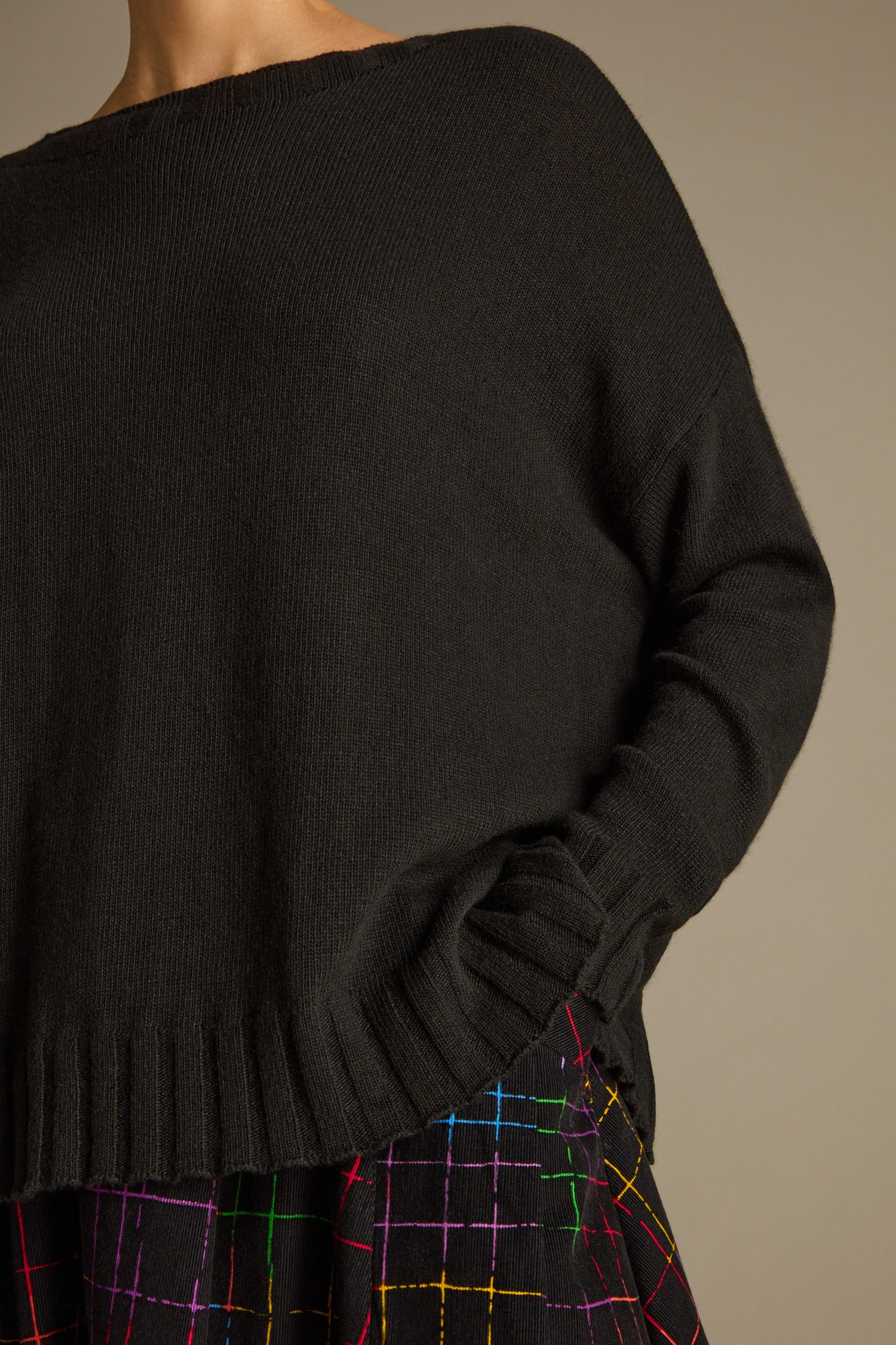 A person is wearing an oversized Merino Blend Crop Jumper paired with a black skirt featuring colorful grid patterns, with one hand in their pocket.