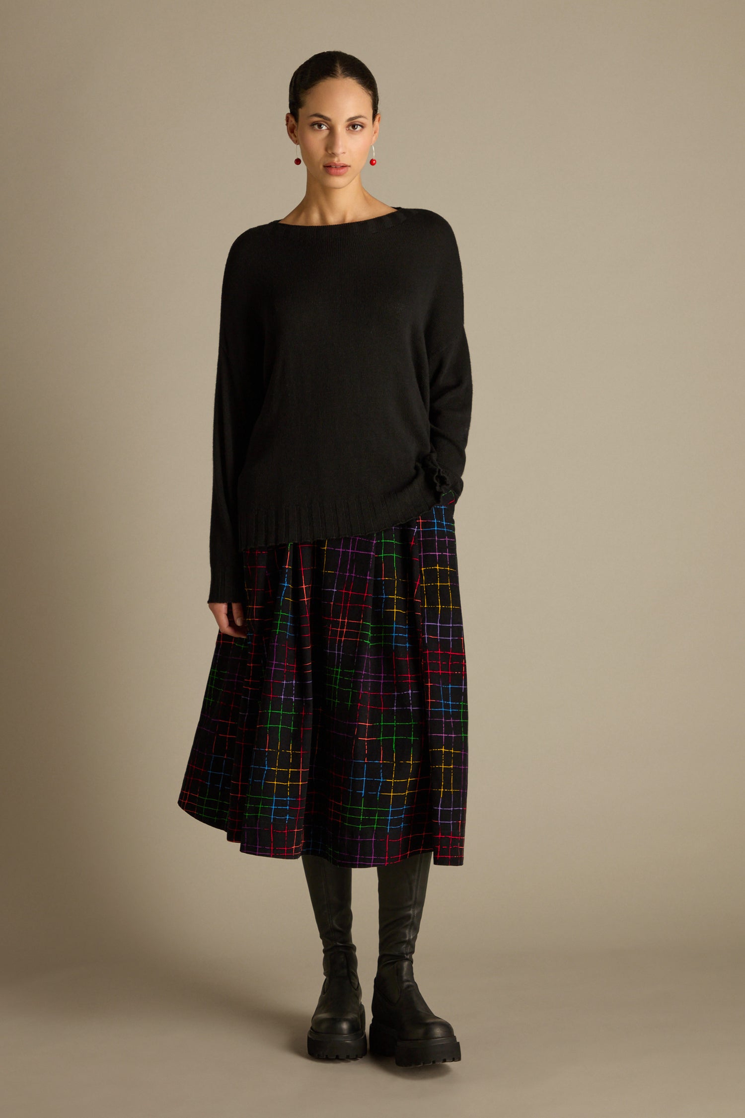 A person stands against a plain background wearing a Merino Blend Crop Jumper, a multicolored checked skirt, and black boots.