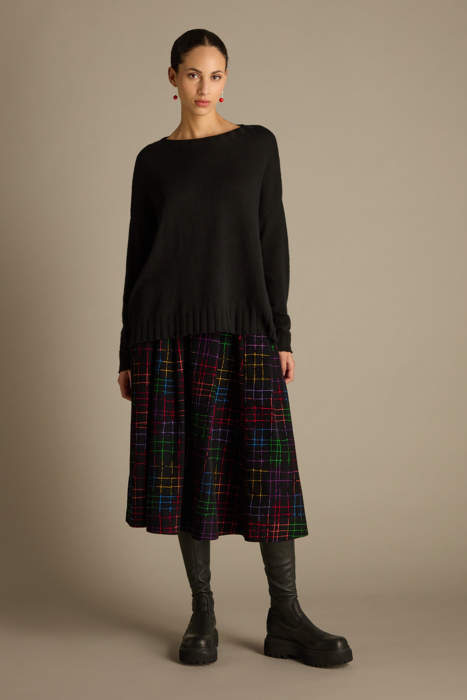 A person stands against a plain background wearing a black Merino Blend Crop Jumper, a colorful plaid skirt, and black boots.