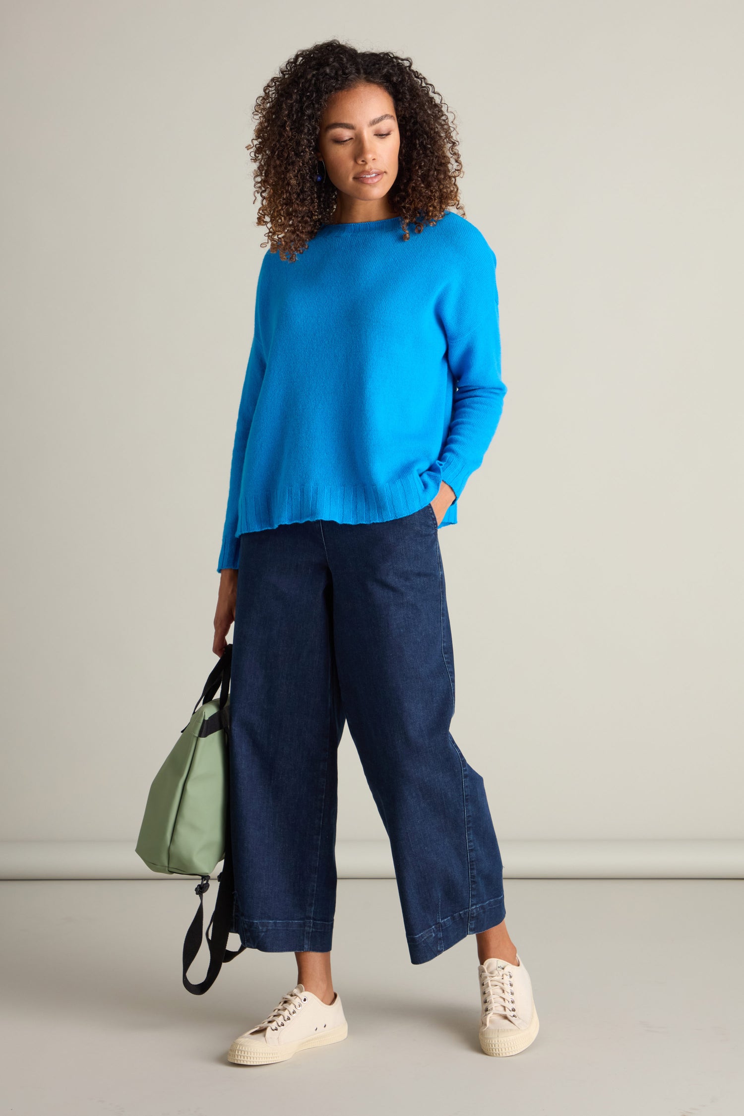 A person with curly hair wearing a Cashmere Blend Crop Jumper, wide-leg dark blue jeans, and cream-colored sneakers stands with a light green bag in hand against a plain background.