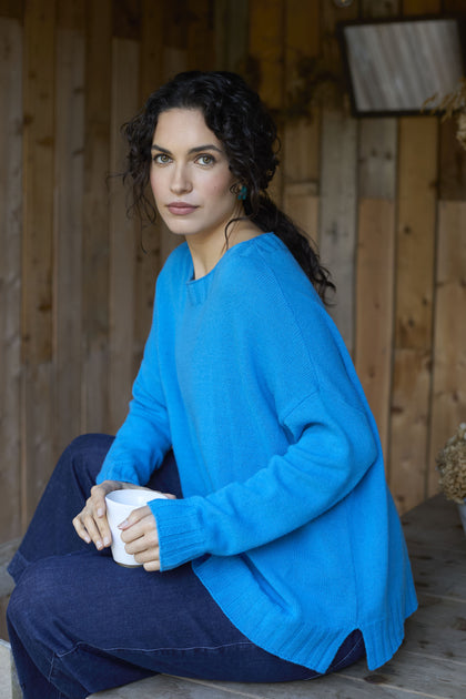 A person with long, dark curly hair wearing a bright blue Cashmere Blend Crop Jumper and dark jeans holds a white mug while sitting against a wooden background.