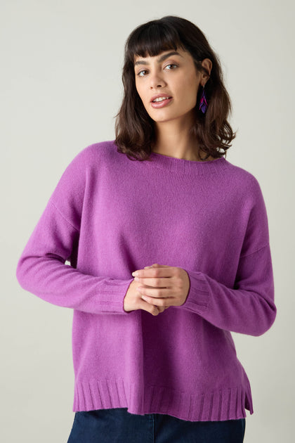 A person with shoulder-length dark hair is wearing a vibrant Merino Wool Crop Jumper and purple earrings, looking towards the camera against a plain background, presenting a striking cropped silhouette.