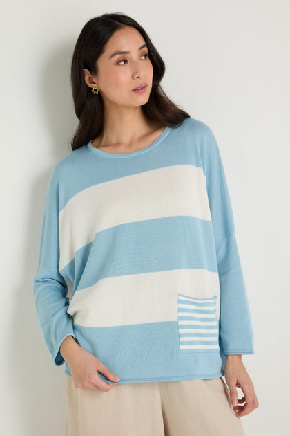 A woman wears the Stripe Pocket Knit Top—a relaxed, boxy knit with white horizontal stripes and a striped pocket—paired with beige pants. Made from soft modal cotton, she poses against a plain light background.