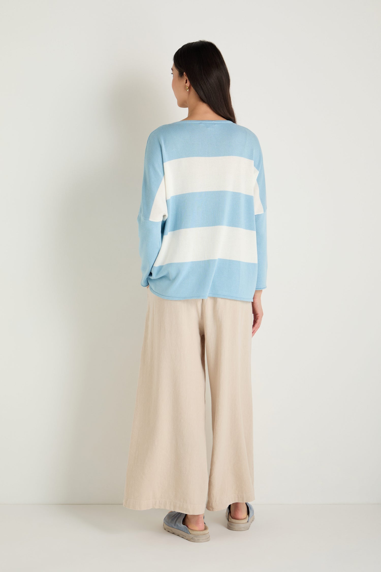 A woman faces away, wearing the Stripe Pocket Knit Top in light blue and white stripes with a relaxed boxy fit, paired with wide-leg beige pants and slip-on shoes against a plain white backdrop.