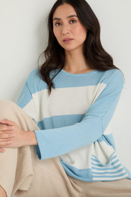 A woman with long dark hair sits against a white wall wearing the Stripe Pocket Knit Top and beige pants, looking at the camera with a neutral expression.