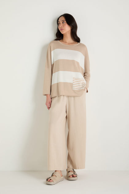 A woman stands against a plain white background wearing the Stripe Pocket Knit Top in a relaxed boxy fit, paired with wide-leg beige pants and beige sandals.