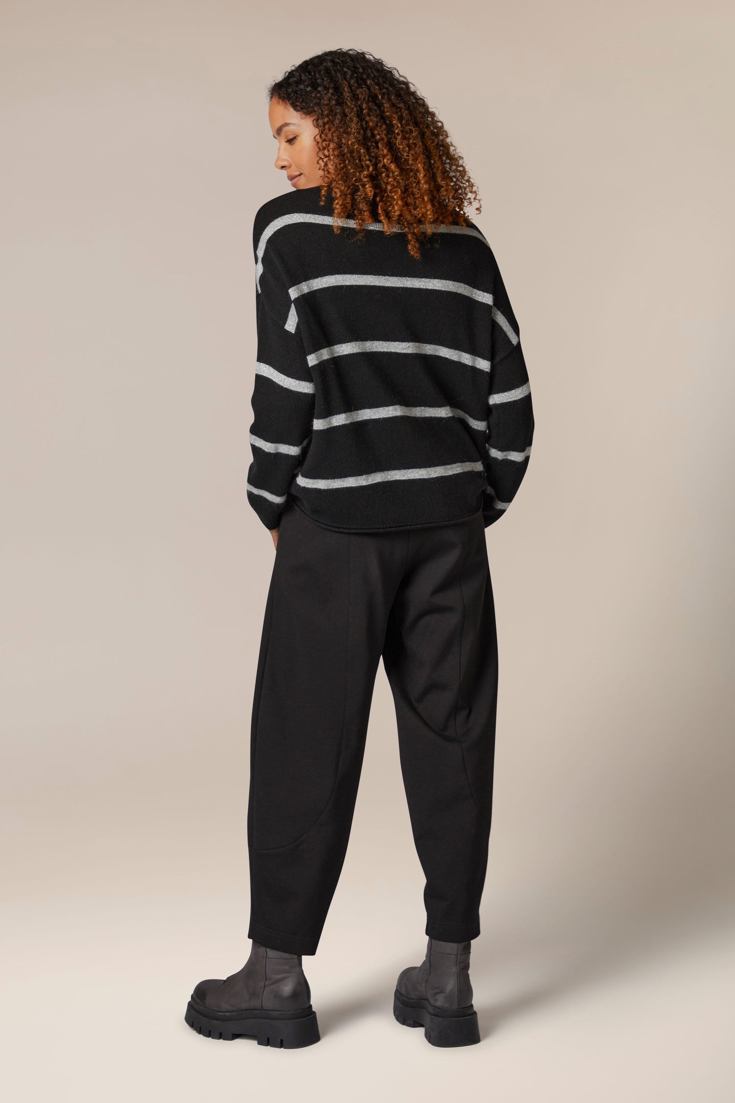 The back view of a woman wearing a Plaited Stripe Sweater with metallic sheen and black trousers.