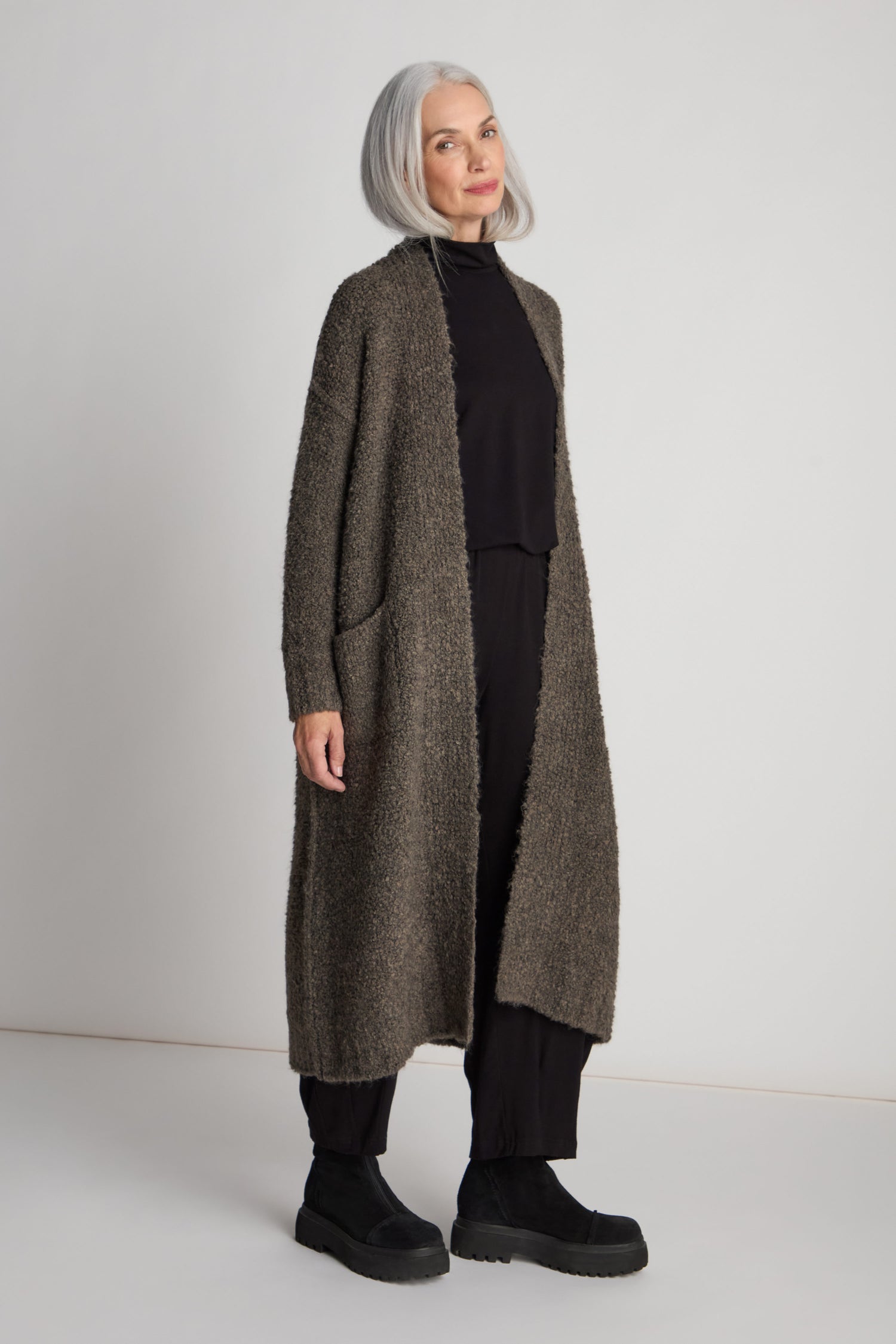 A woman with gray hair wears the Boucle Knit Long Cardi over a black outfit and black boots, standing against a plain light background. This layering essential creates an elegant long-line silhouette.