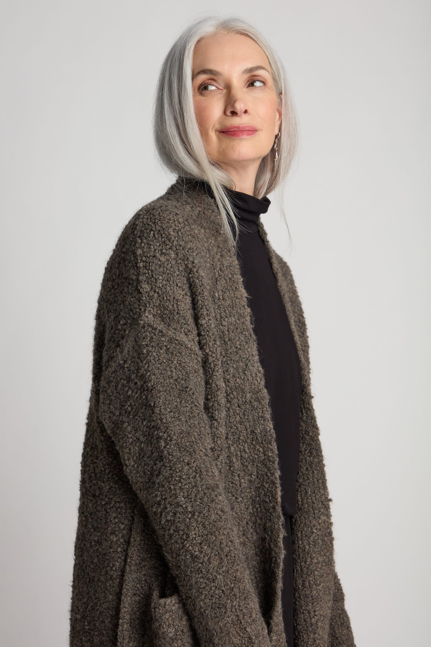 A person with long grey hair, wearing a dark turtleneck and the Boucle Knit Long Cardi in a textured brown finish, looks off to the side against a plain background. The ensemble features a long-line silhouette that serves as the perfect layering essential.