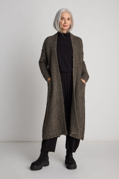 An older adult with shoulder-length gray hair stands against a plain background, wearing a long-line silhouette with the Boucle Knit Long Cardi in dark colors, paired with a black turtleneck, black pants, and black boots. Hands are in the cardigan pockets.