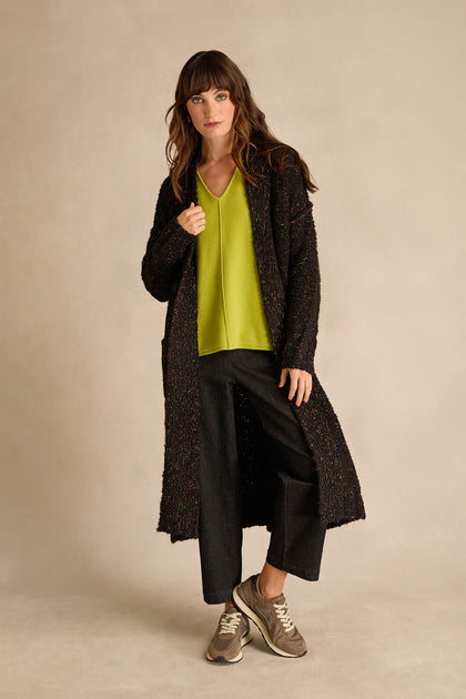 A woman stands in a studio against a neutral background, wearing the Black Boucle Knit Long Line Speckle Cardi layered over a lime green V-neck top, paired with black pants and beige sneakers.