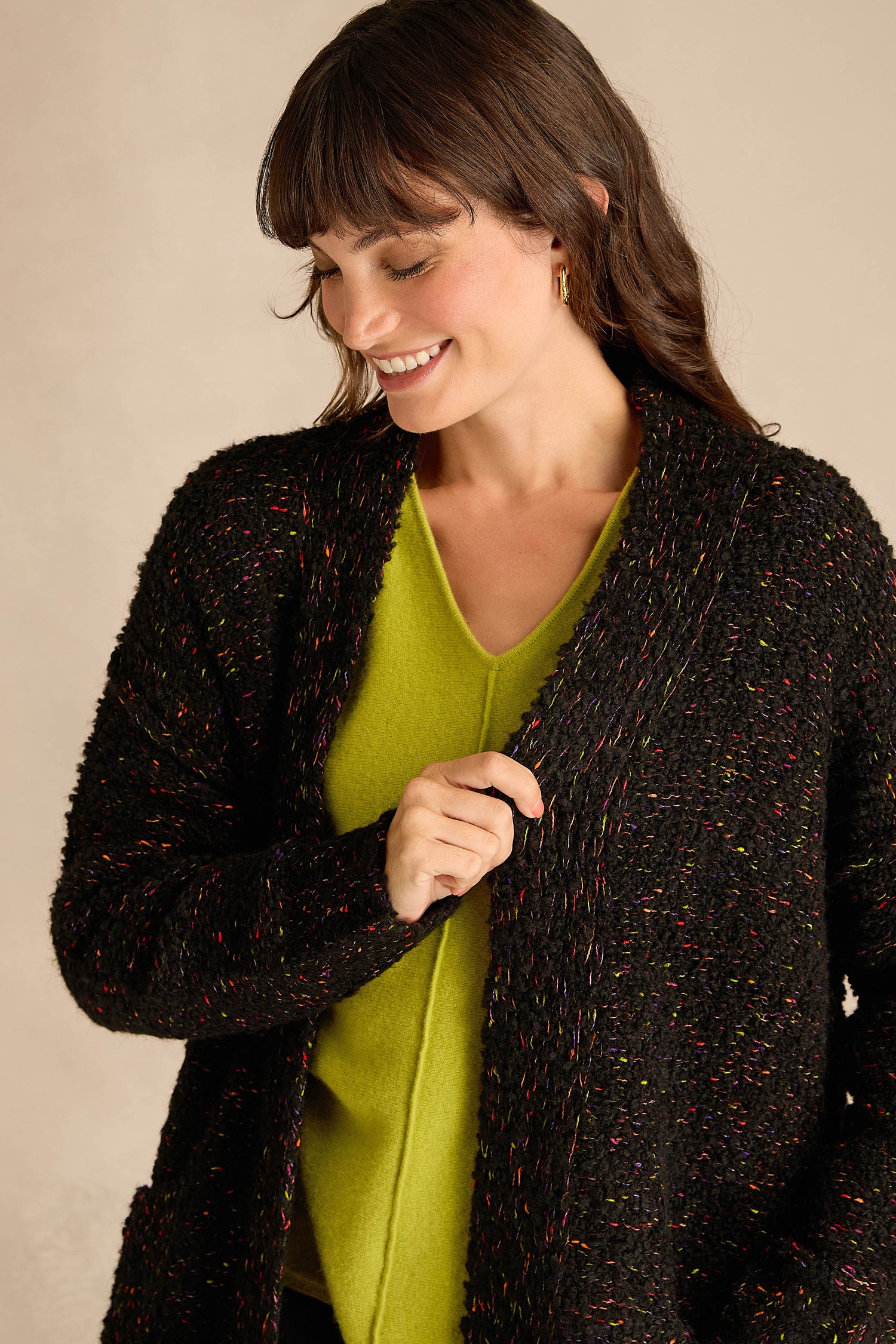 A woman with dark hair wears a bright green V-neck top and the Black Boucle Knit Long Line Speckle Cardi while looking down and smiling.