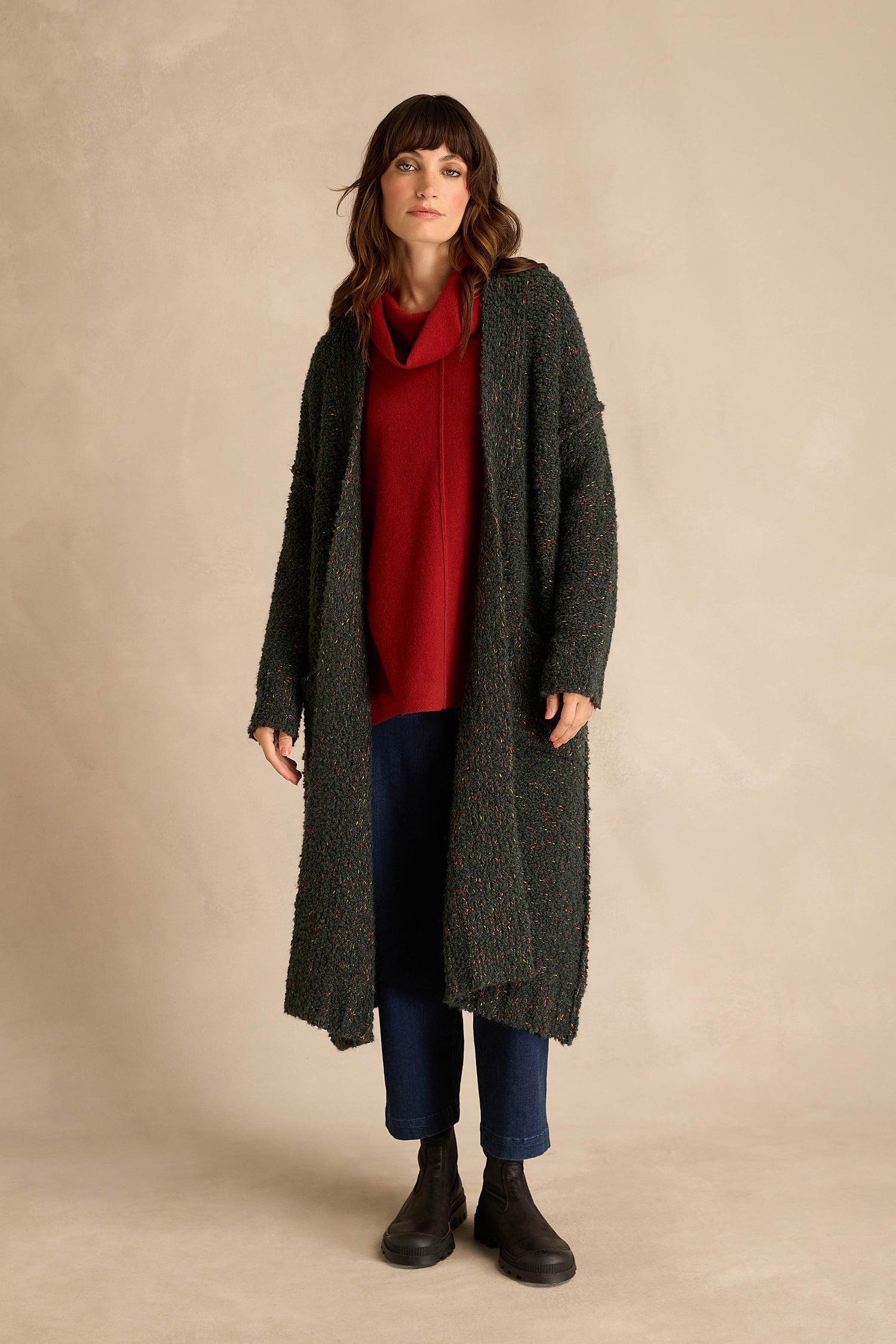 A woman stands against a beige background, wearing the Forest Boucle Knit Long Line Speckle Cardi over a red cowl neck top, blue pants, and black boots—a stylish take on modern women’s knitwear.