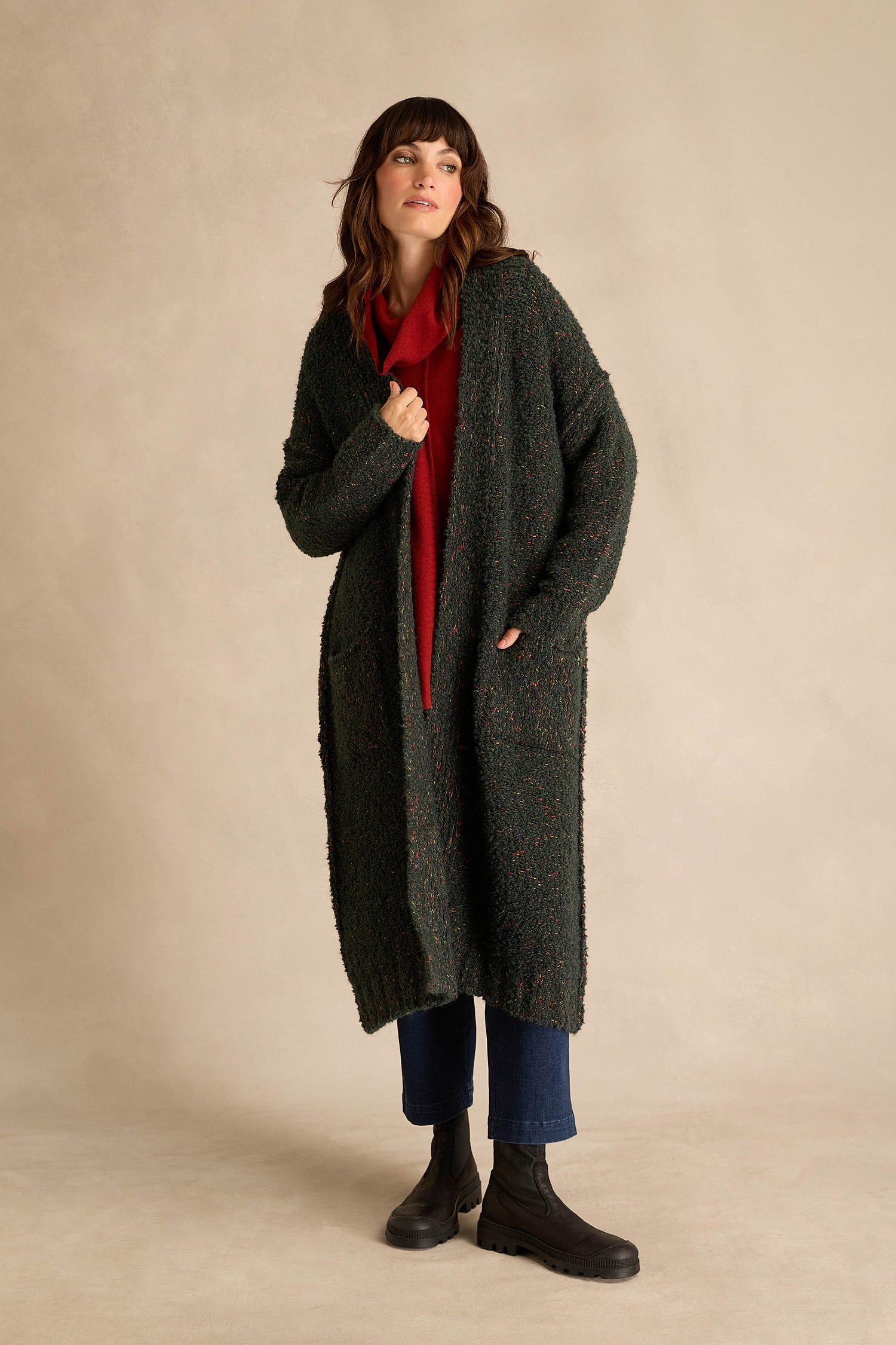 A person wearing the Forest Boucle Knit Long Line Speckle Cardi—a long, dark green textured cardigan—paired with a red top, blue jeans, and black boots stands with one hand in a pocket against a plain beige background.