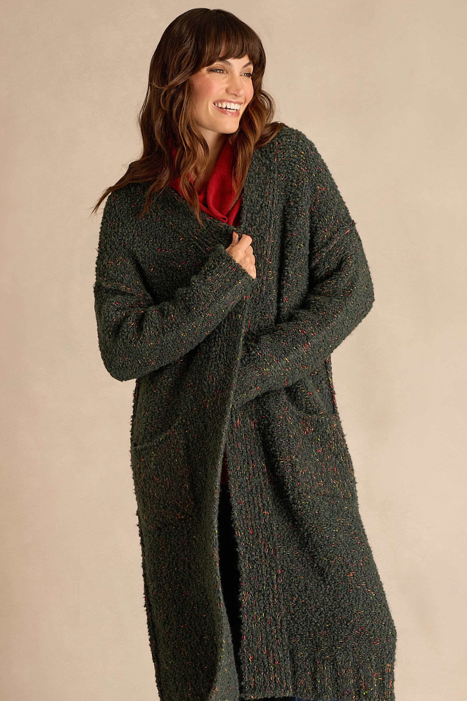 A woman stands and smiles against a plain, light-colored background, wearing the Forest Boucle Knit Long Line Speckle Cardi—a long, textured dark green cardigan—layered over a red top.