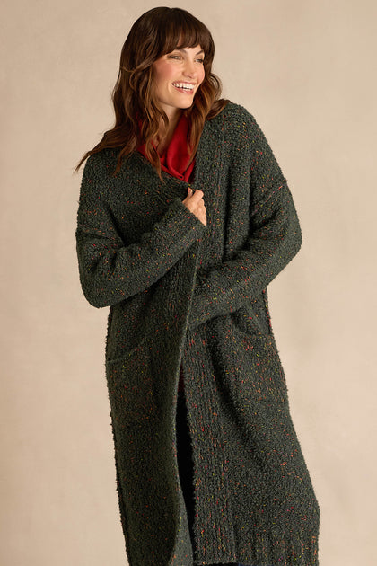 A woman stands and smiles against a plain, light-colored background, wearing the Forest Boucle Knit Long Line Speckle Cardi—a long, textured dark green cardigan—layered over a red top.