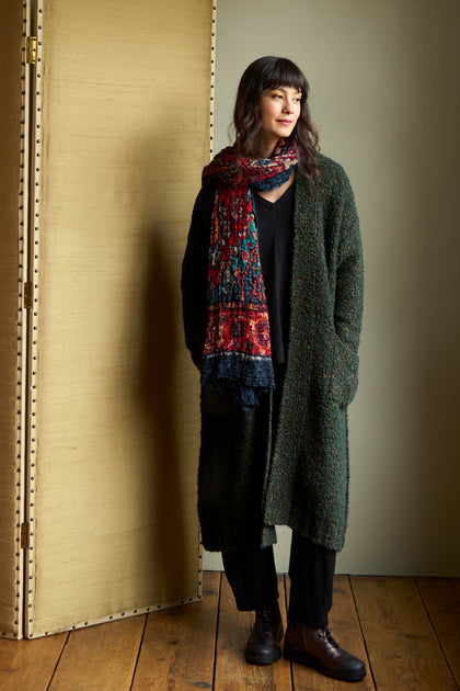 A woman wearing a Forest Boucle Knit Long Line Speckle Cardi over a black top and trousers, styled with a colorful patterned scarf and dark boots.