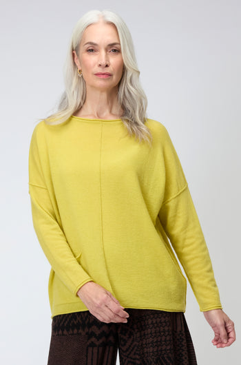 An older woman with long gray hair stands facing the camera against a plain background, wearing a yellow Merino Pocket Jumper and dark patterned pants.