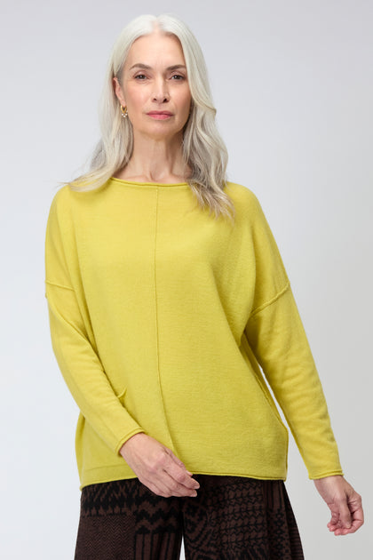 An older woman with long gray hair stands facing the camera against a plain background, wearing a yellow Merino Pocket Jumper and dark patterned pants.