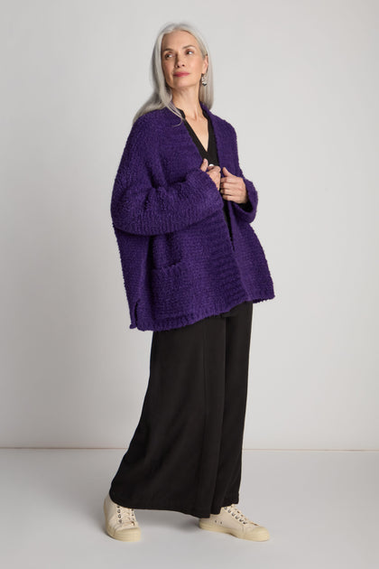 A person with long gray hair wearing a Boucle Knit Cable Trim Cardi in purple, black pants, and white shoes stands against a plain background.