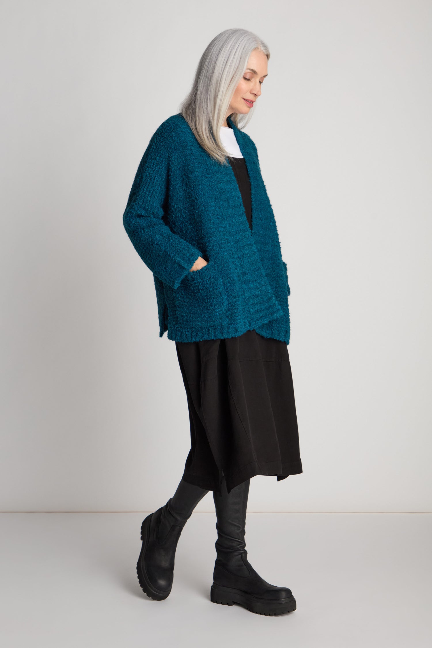 A woman with long gray hair is standing sideways, her hands tucked into the pockets of her teal Boucle Knit Cable Trim Cardi. She pairs it with a black dress, black leggings, and black boots.