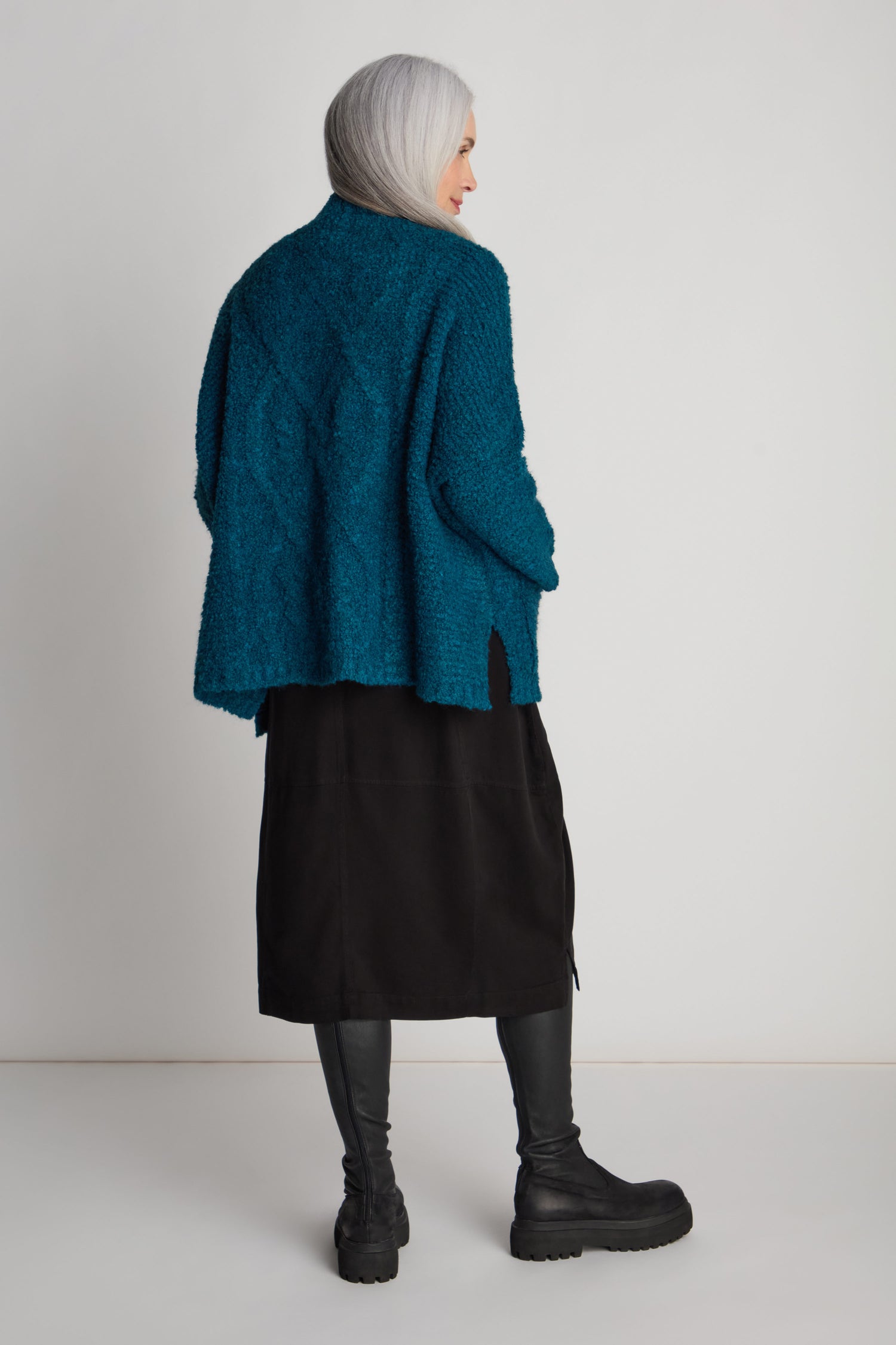 A person with long gray hair is wearing a teal Boucle Knit Cable Trim Cardi, a black skirt, black leggings, and black boots, and is standing with their back to the camera.