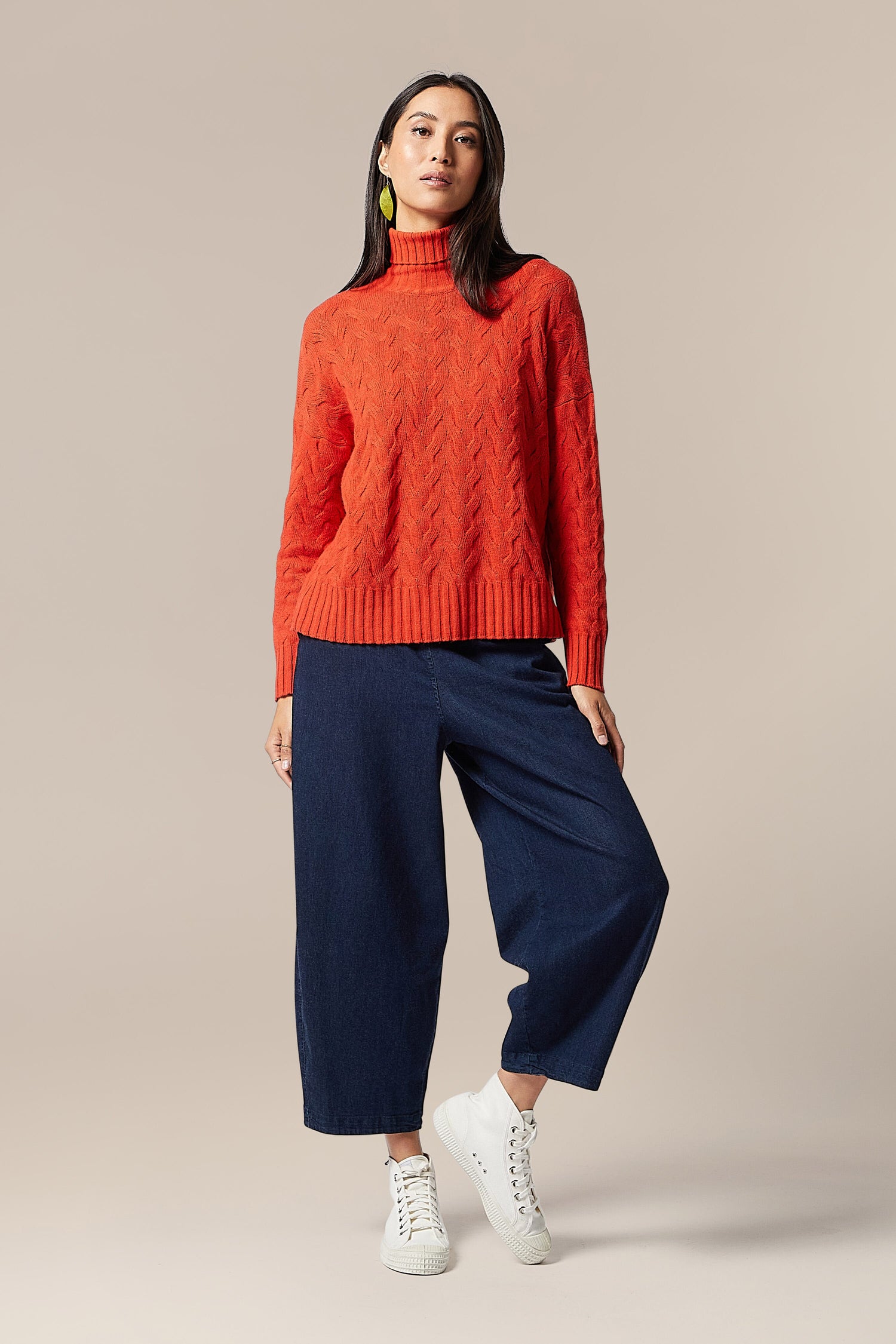A woman wearing a high-quality Cable Knit Jumper in an attractive orange hue paired with fashionable blue wide leg pants.