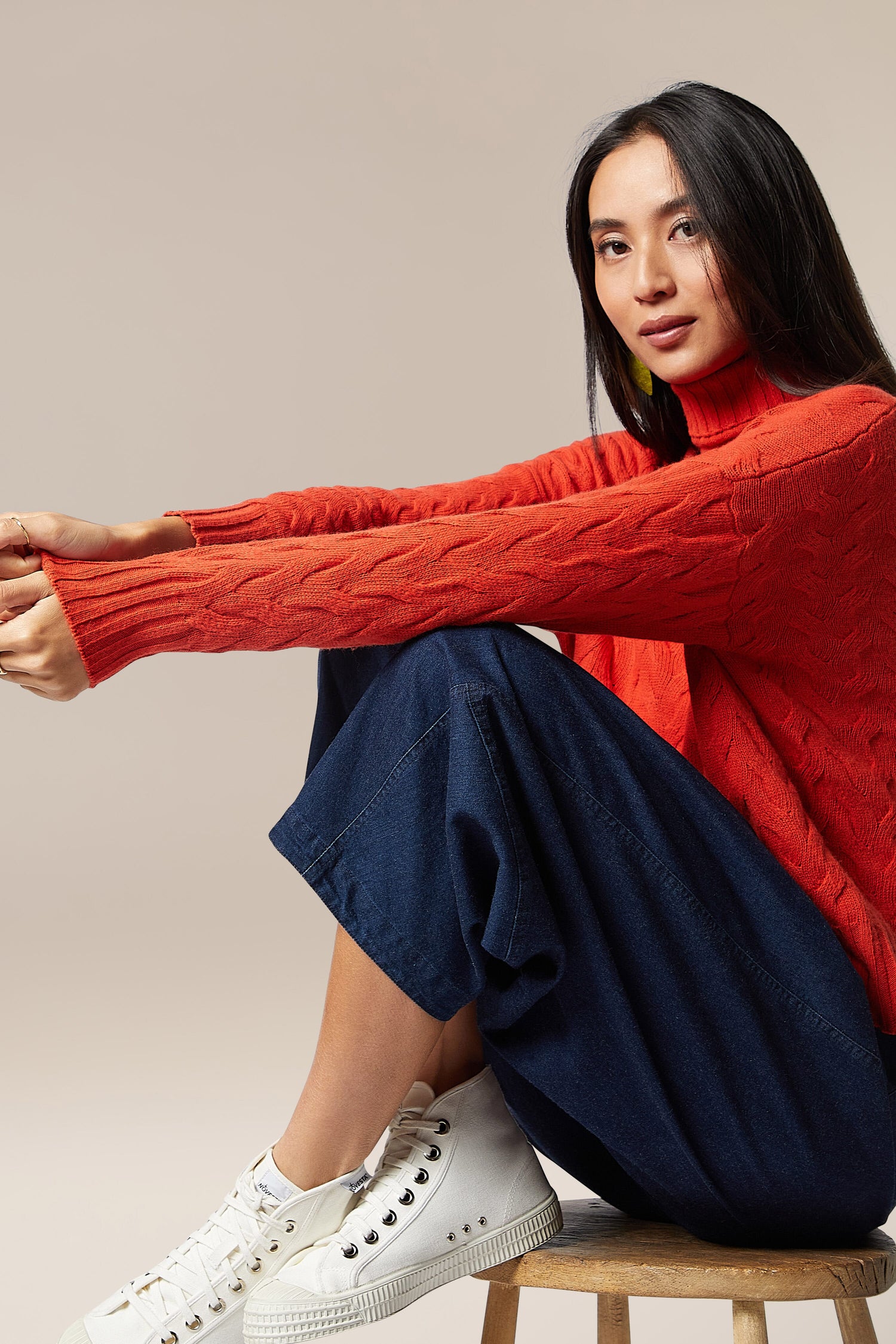 A woman in a cozy Cable Knit Jumper, made of quality merino wool, sitting on a stool.