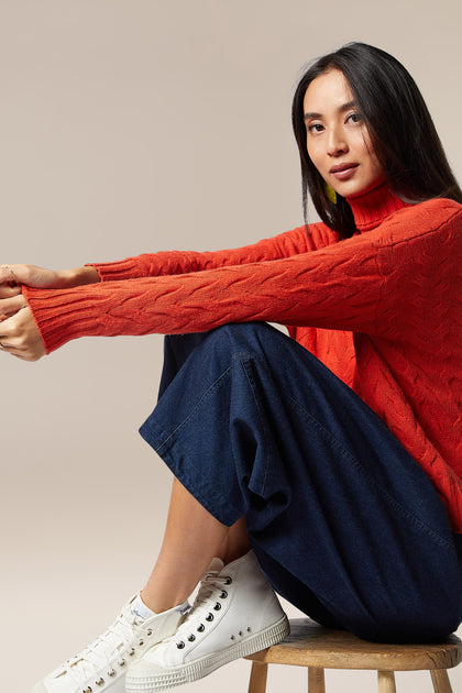 A woman in a cozy Cable Knit Jumper, made of quality merino wool, sitting on a stool.