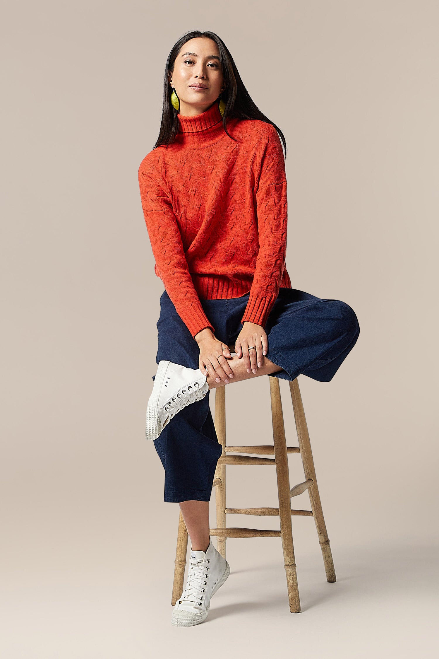 A woman in a high-quality knitwear, a Cable Knit Jumper, sitting on a stool.