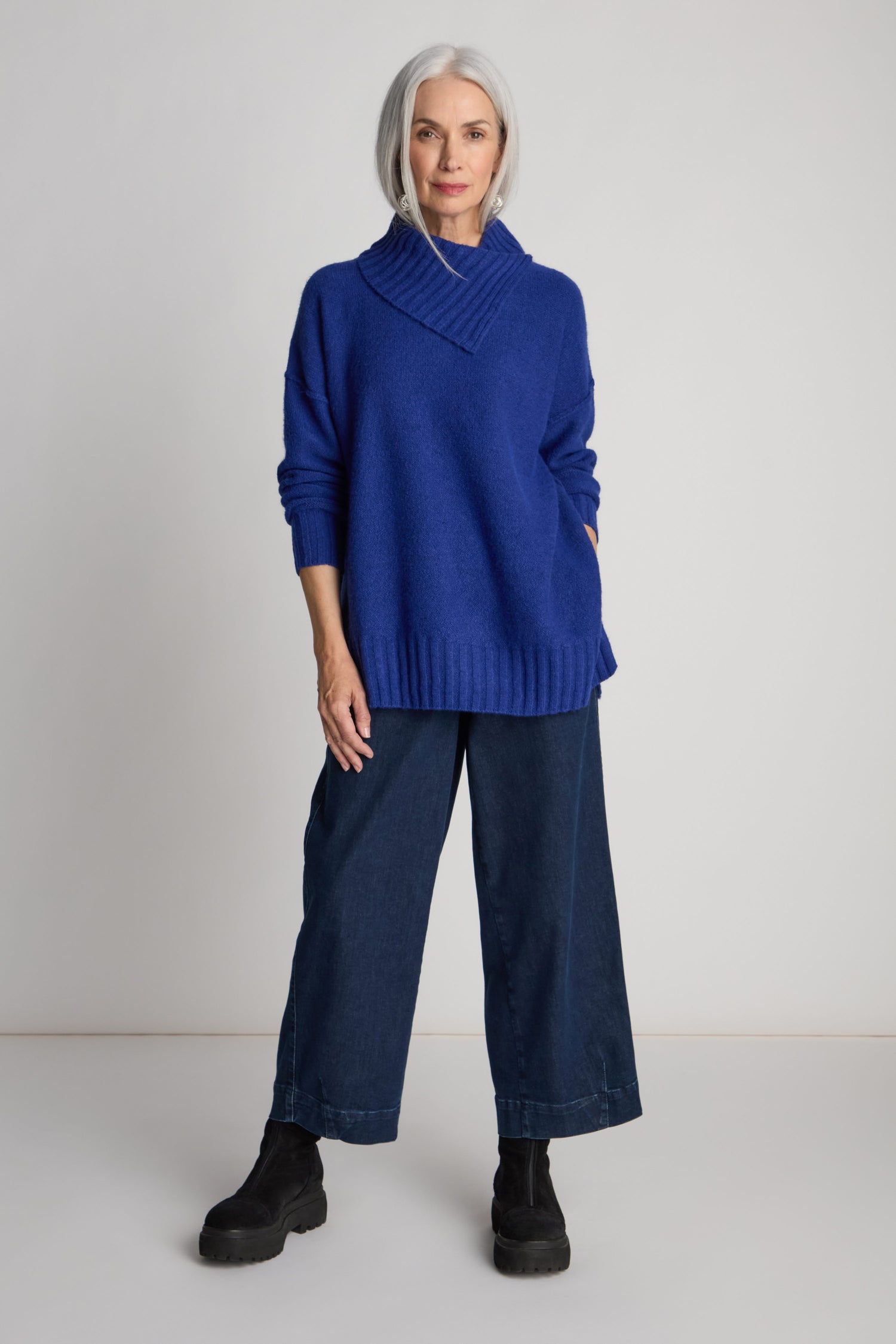 A woman with shoulder-length gray hair is wearing a blue Wool Viscose Mix Asymmetric Jumper in a contemporary style, paired with blue wide-leg pants and black boots. She stands against a plain white background.