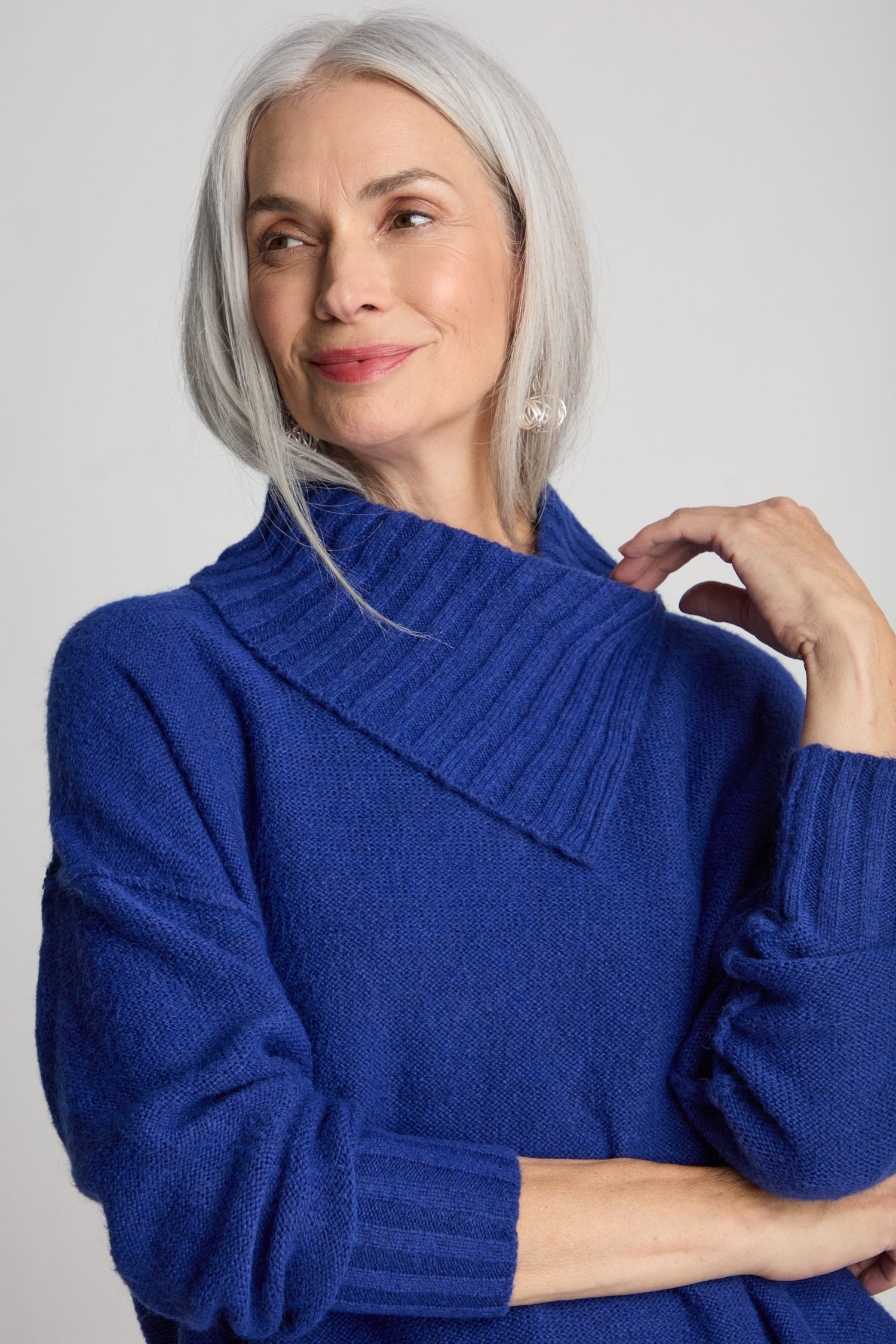 A woman with shoulder-length gray hair, smiling and touching her collar with one hand, is wearing a Wool Viscose Mix Asymmetric Jumper in a contemporary style.