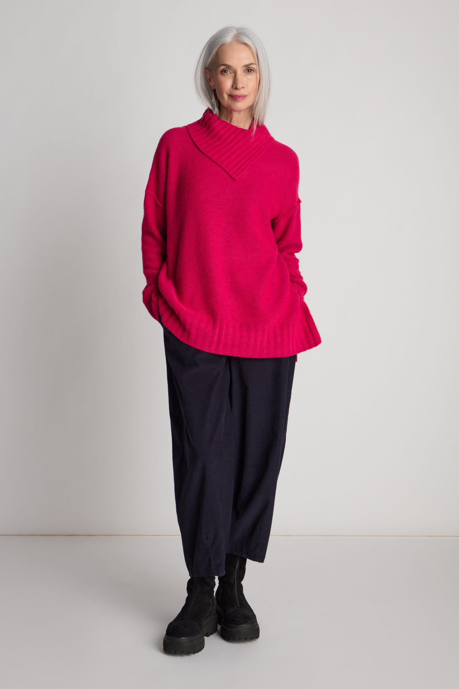 A woman with gray hair stands against a plain background, clad in a bright pink Wool Viscose Mix Asymmetric Jumper and black pants, paired with black boots. Her hands rest casually in her sweater pockets, exuding contemporary style.