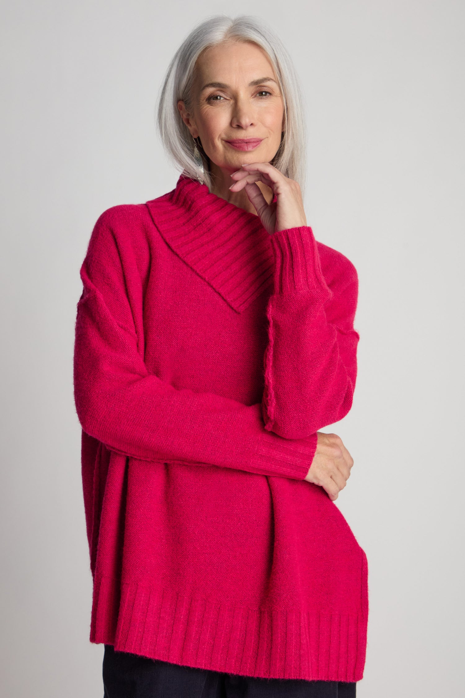 A person with long gray hair poses confidently, wearing a contemporary-style bright pink Wool Viscose Mix Asymmetric Jumper with a high neckline.