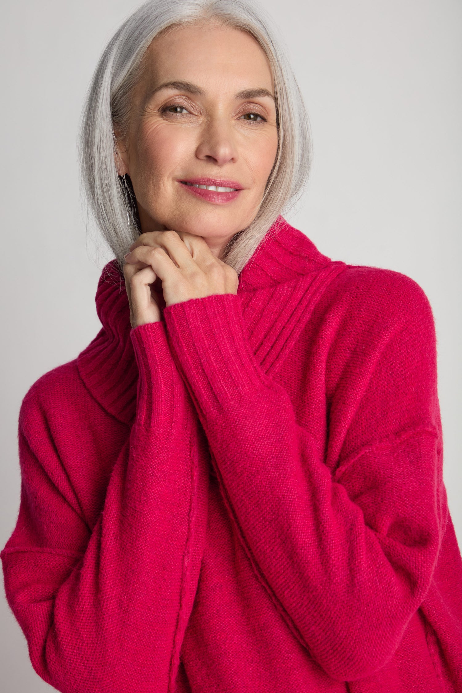 A woman with long gray hair and a gentle smile wearing a bright pink Wool Viscose Mix Asymmetric Jumper with a ribbed high collar stands against a plain light-colored background.