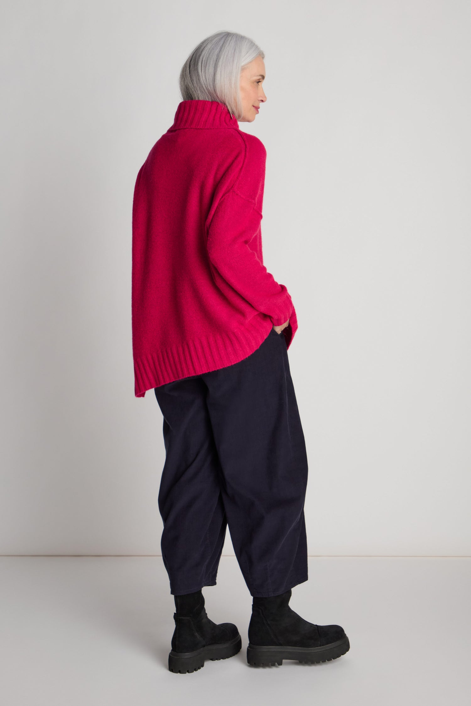 A person with shoulder-length gray hair is wearing a bright pink Wool Viscose Mix Asymmetric Jumper, dark blue pants, and black boots. They are standing against a plain white background and facing to the side.