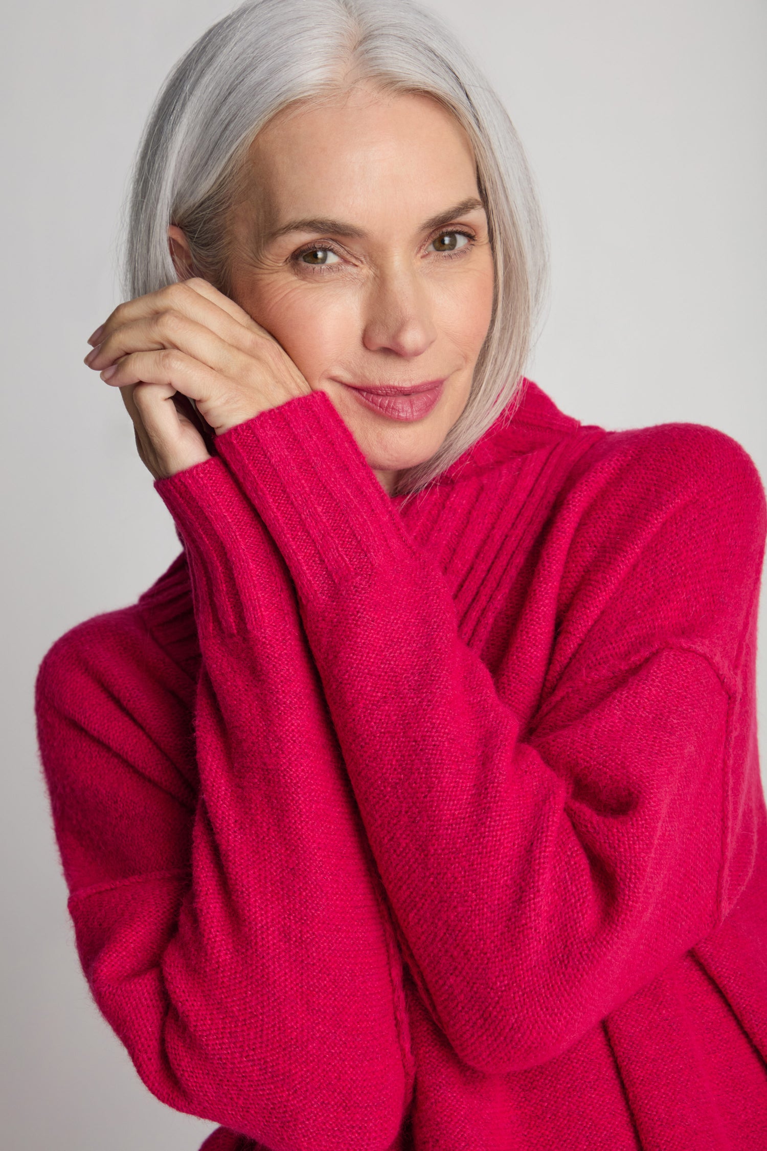 A woman with gray hair is wearing the bright pink Wool Viscose Mix Asymmetric Jumper. She lightly rests her hands near her face and looks at the camera against a plain background, embodying contemporary style.