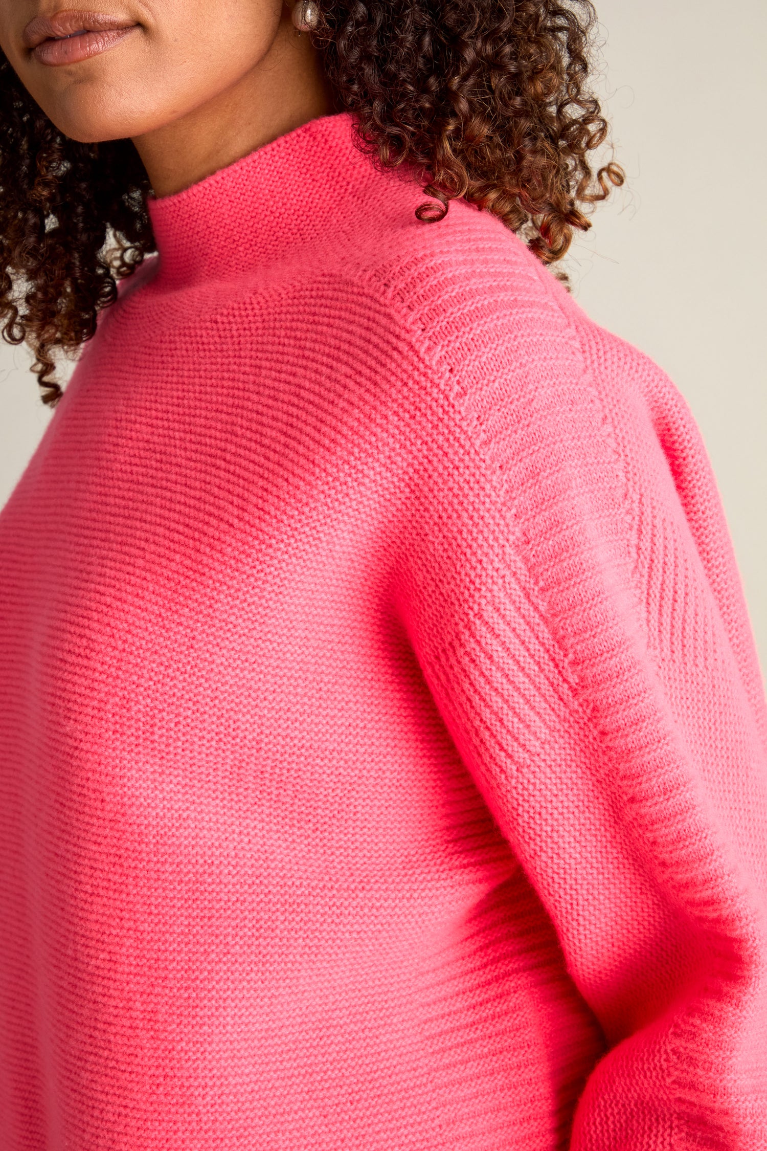 A person wearing a bright pink Merino Wool Stitch Detail Jumper with curly hair visible on the left side.