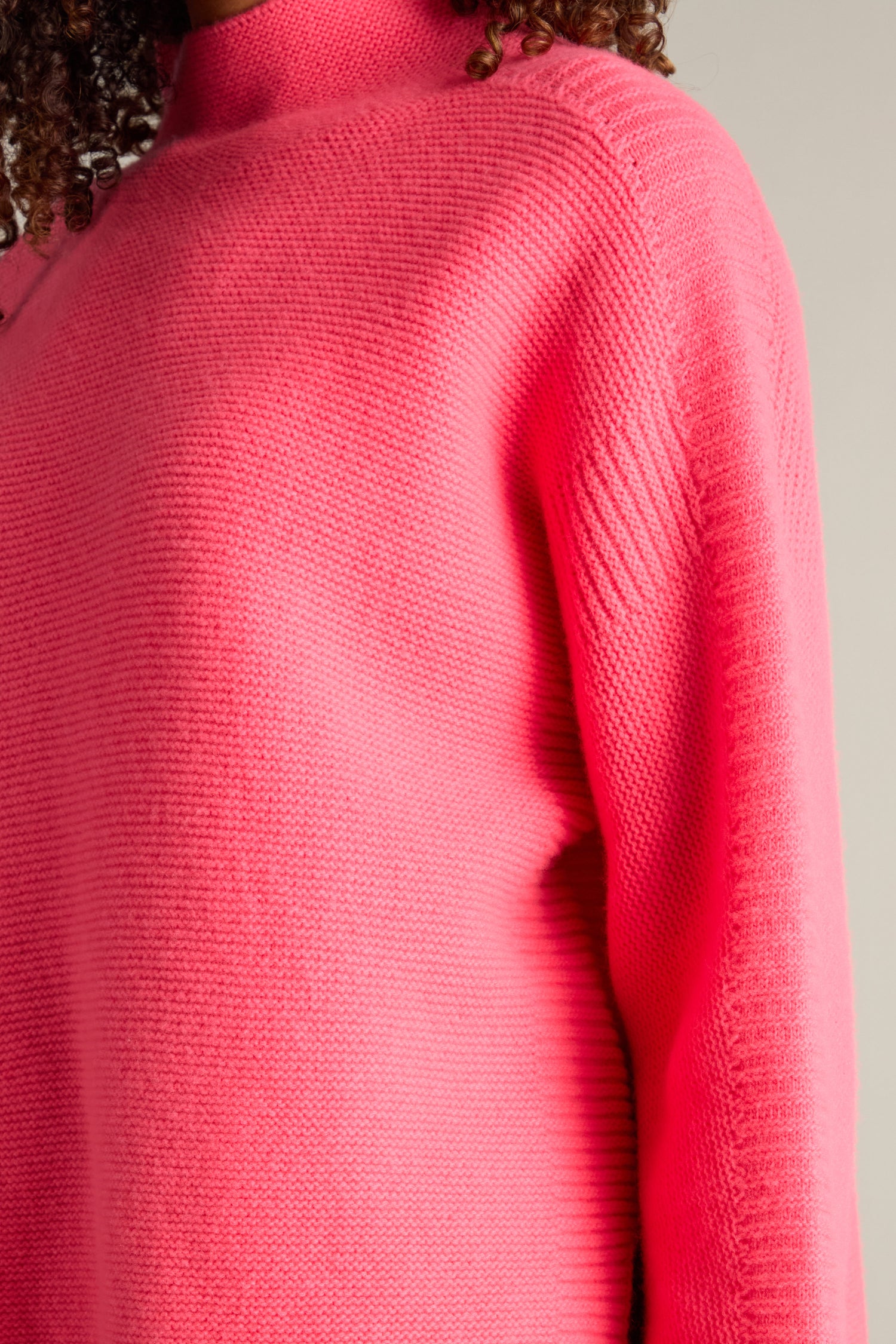 Close-up of a person wearing a Merino Wool Stitch Detail Jumper in bright pink, with visible curly hair on one side.