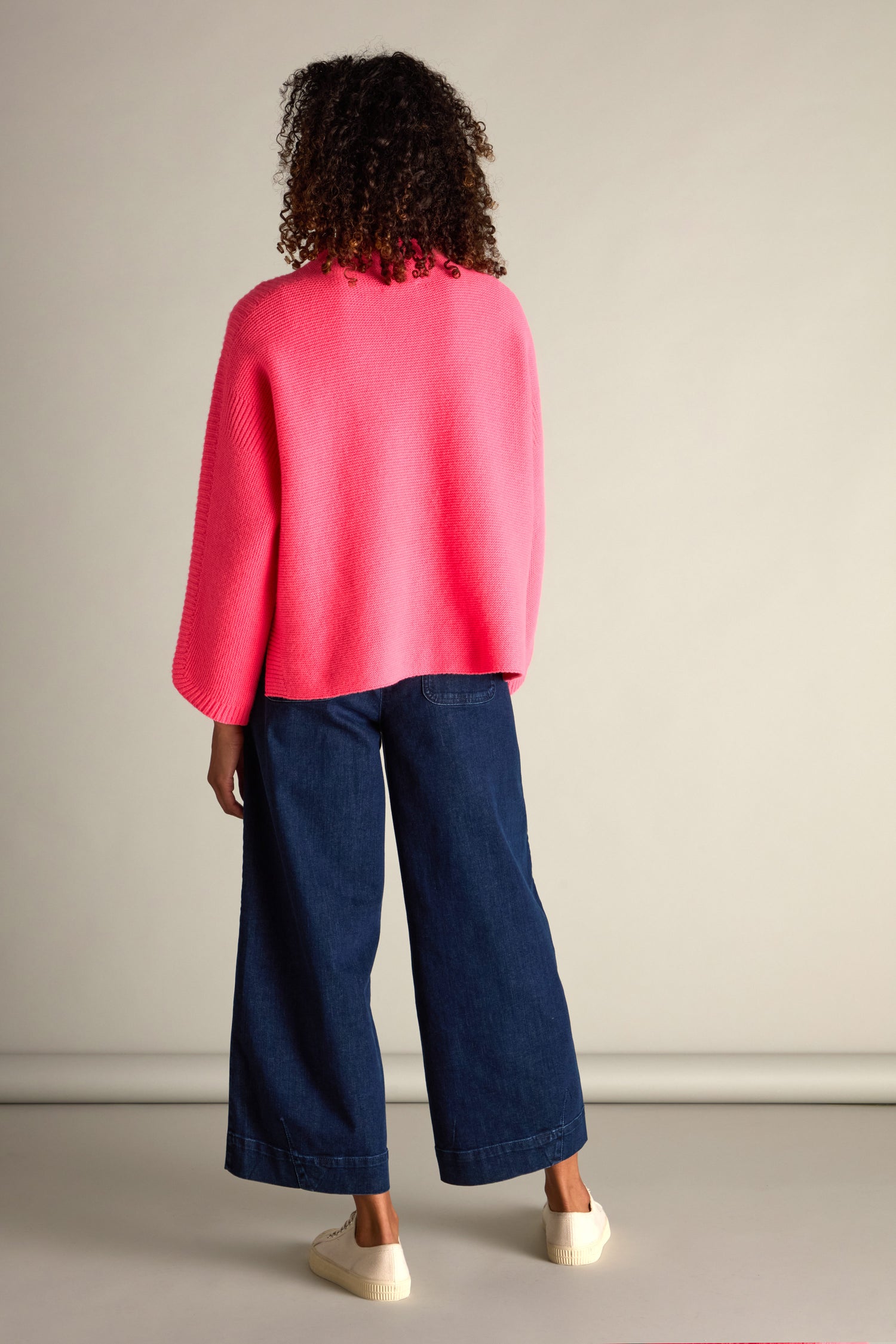 The person with curly hair stands facing away, wearing a bright pink Merino Wool Stitch Detail Jumper, blue wide-legged jeans, and white sneakers, exuding cozy comfort against a neutral background.