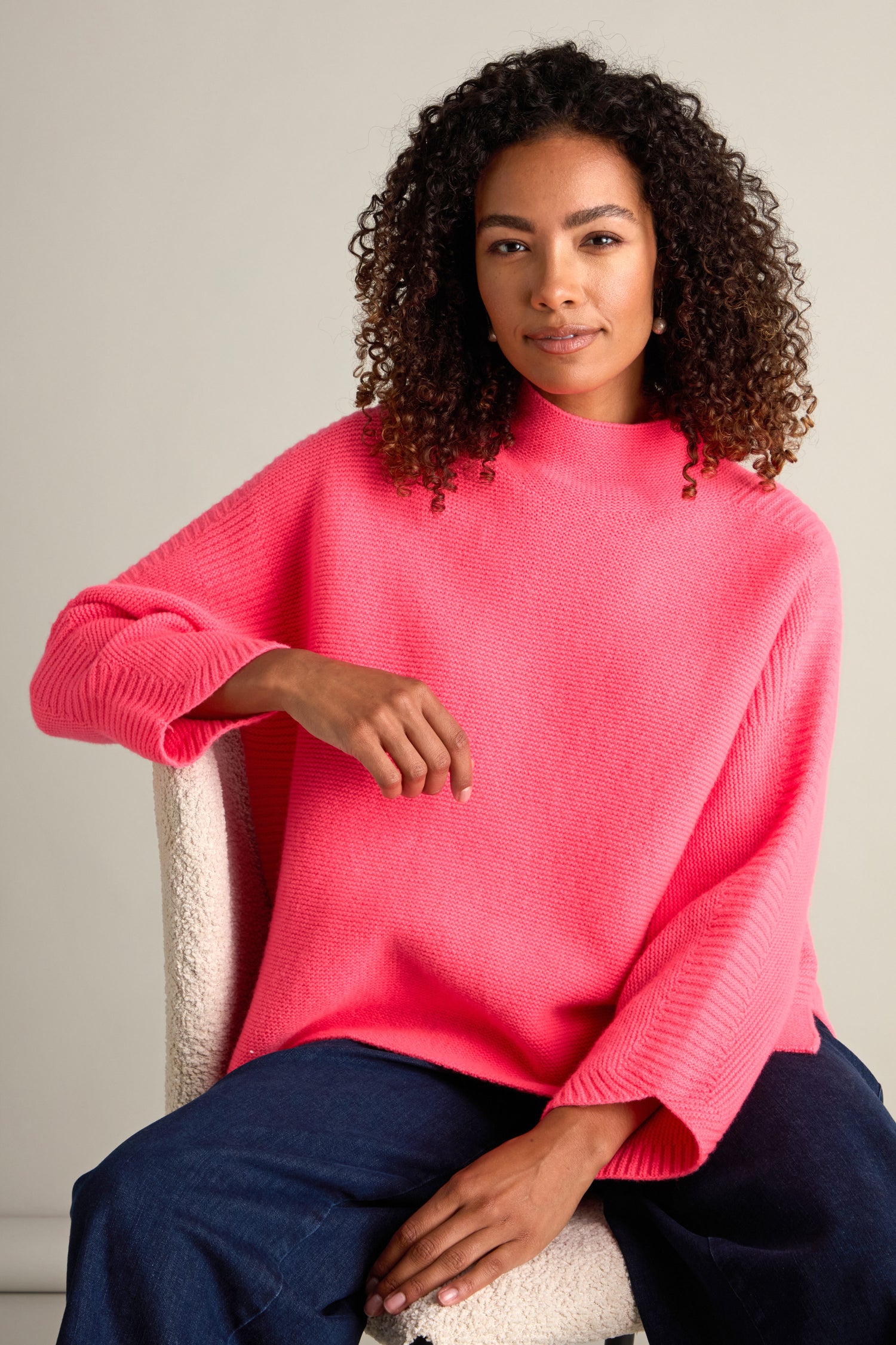 A person with curly hair is seated, wearing a bright pink Merino Wool Stitch Detail Jumper and blue pants. They are looking forward with a neutral expression, embodying cozy comfort.