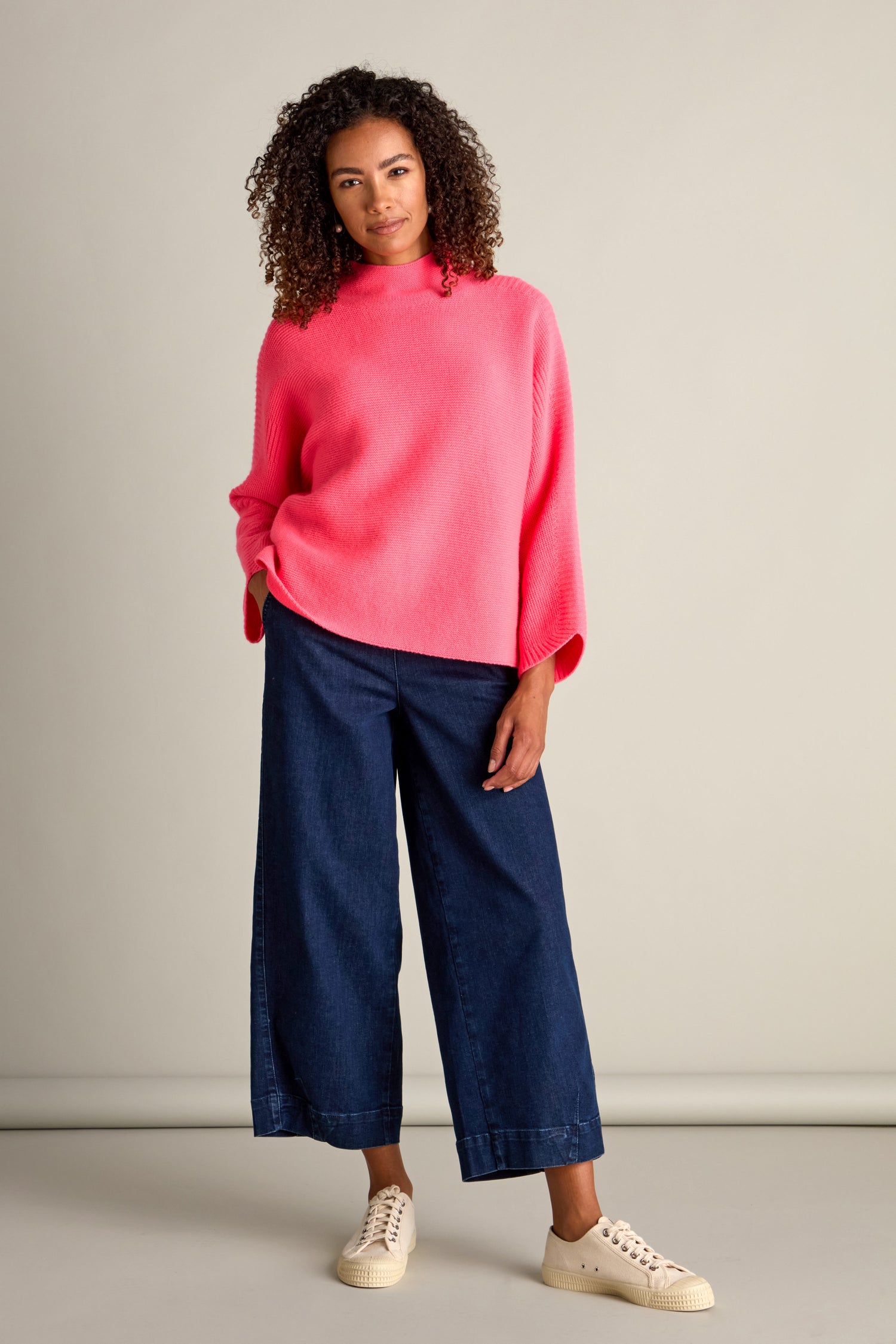 A person stands against a plain background, wearing a bright pink Merino Wool Stitch Detail Jumper, blue wide-leg jeans, and white sneakers. Embracing cozy comfort, the person's left hand is in their pocket.