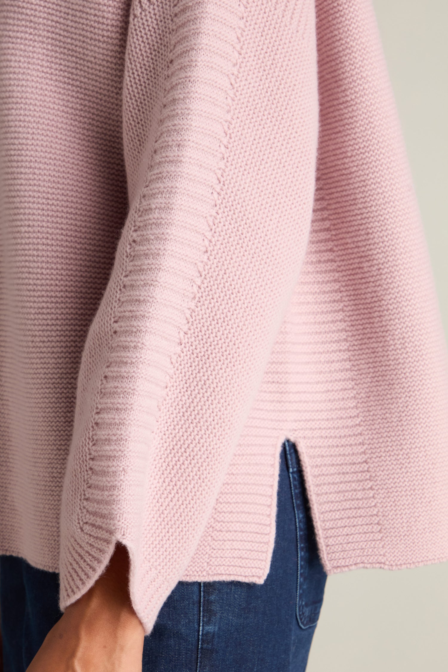 A close-up of a person wearing the light pink, textured Merino Wool Stitch Detail Jumper with a side slit, paired with dark blue jeans. The image highlights the cozy comfort of the knitwear and focuses on the person's hip area.