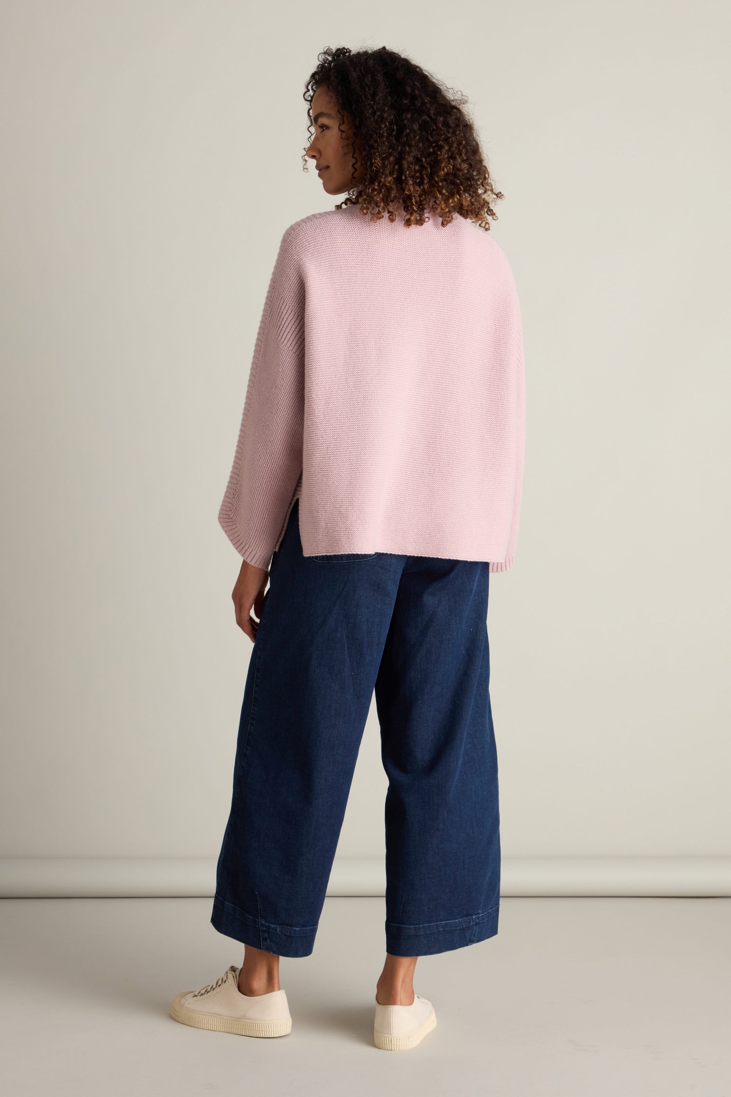 A person with curly hair is standing and facing away, wearing a light pink Merino Wool Stitch Detail Jumper, dark blue wide-leg jeans, and white sneakers against a plain background.