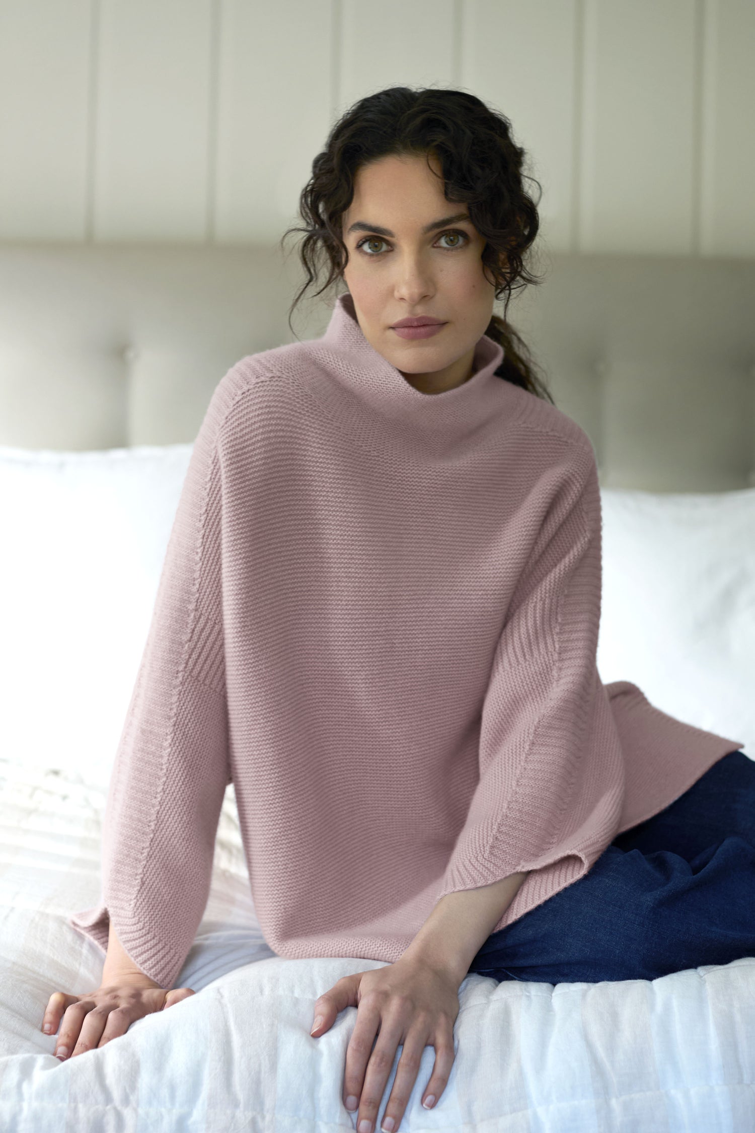 A person with dark curly hair sits on a bed wearing a light pink Merino Wool Stitch Detail Jumper and blue pants, gazing directly at the camera.